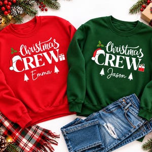 May include: A red sweatshirt and a dark green sweatshirt, each featuring the white text "Christmas CREW" with a Santa hat, a gift box, and two pine trees. The red sweatshirt has the name "Emma" and the green sweatshirt has the name "Jason".