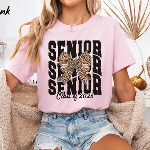 Puede incluir: Camiseta rosa con un gr&aacute;fico en blanco y negro que dice "Senior" con un lazo de estampado de leopardo en el medio y "Class of 2026" en la parte inferior.
