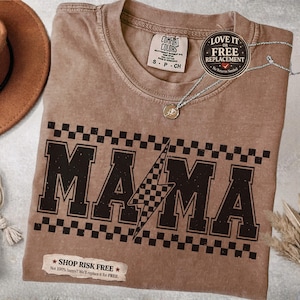 May include: A brown Comfort Colors t-shirt with the word "MAMA" in a bold, black font, accented with a checkered pattern and a lightning bolt. The shirt has a small tag that says "S-P-CH". A silver necklace with a pendant is on the shirt.
