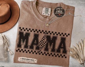 May include: A brown Comfort Colors t-shirt with the word "MAMA" in a bold, black font, accented with a checkered pattern and a lightning bolt. The shirt has a small tag that says "S-P-CH". A silver necklace with a pendant is on the shirt.