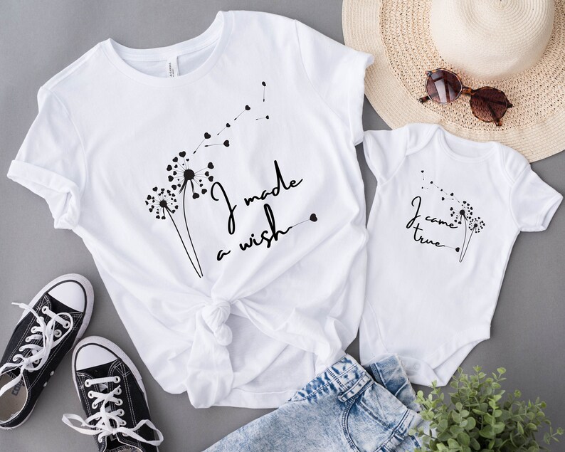 Op de afbeelding: Wit T-shirt met een paardenbloemdessin en de tekst "Ik heb een wens gedaan". Een wit rompertje met een paardenbloemdessin en de tekst "Ik ben uitgekomen".