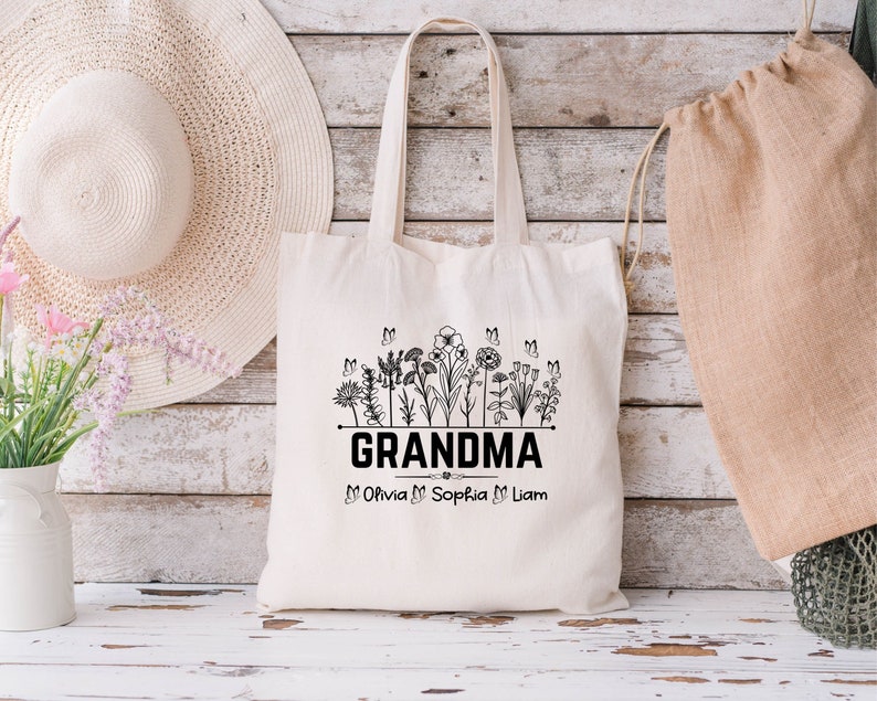 Op de afbeelding: Een witte canvas tote bag met zwarte tekst die "Grandma" zegt en een rij bloemen en vlinders. Onder de tekst staan de namen "Olivia, Sophia, Liam".