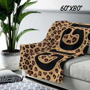 K&ouml;nnte beinhalten: Eine braune und schwarze Leopardenfell-Decke mit schwarzen abstrakten Formen. Die Decke ist 152,4 cm x 203,2 cm gro&szlig; und liegt auf einem Sofa.
