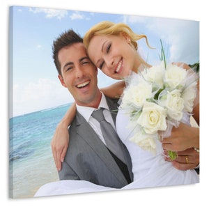 Puede incluir: Impresi&oacute;n en lienzo de una pareja sonriente en una playa. El hombre lleva un traje gris y corbata, mientras que la mujer sostiene un ramo de rosas blancas y lleva un vestido de novia blanco. El fondo muestra el oc&eacute;ano y el cielo azul.
