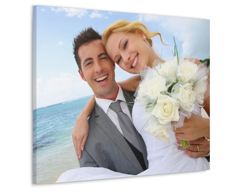 Puede incluir: Impresi&oacute;n en lienzo de una pareja sonriente en una playa. El hombre lleva un traje gris y corbata, mientras que la mujer sostiene un ramo de rosas blancas y lleva un vestido de novia blanco. El fondo muestra el oc&eacute;ano y el cielo azul.