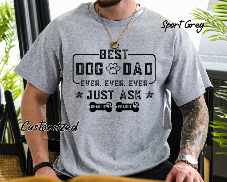 May include: A light gray t-shirt with the text "BEST DOG DAD EVER, EVER, EVER JUST ASK" in black. The design includes a paw print and dog bone graphics, with the names "CHARLIE" and "PEANUT" below. The shirt is labeled "Sport Grey" and "Customized".