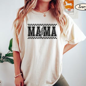 May include: Ivory-colored t-shirt with the word "MAMA" in bold, black letters. A checkered pattern borders the text, and a lightning bolt graphic is in the center. The shirt is a relaxed fit.