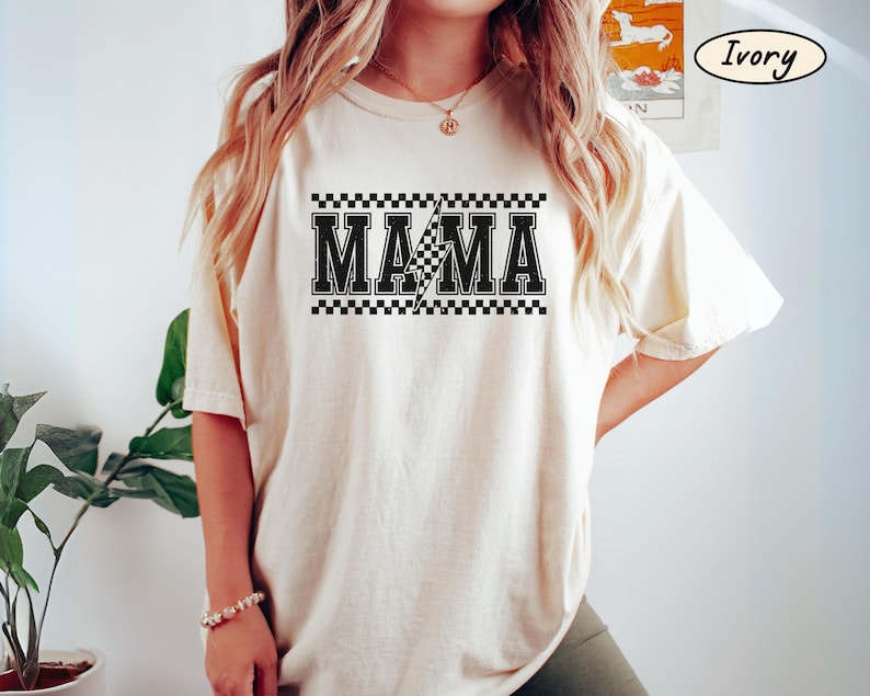 May include: Ivory-colored t-shirt with the word "MAMA" in bold, black letters. A checkered pattern borders the text, and a lightning bolt graphic is in the center. The shirt is a relaxed fit.