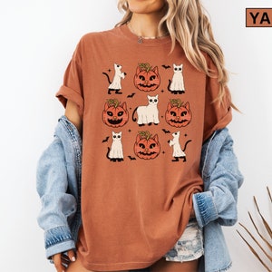 K&ouml;nnte beinhalten: Ein rostorangefarbenes T-Shirt mit einem sich wiederholenden Muster von Halloween-Illustrationen. Das Design umfasst K&uuml;rbislaternen, Geister, Flederm&auml;use und schwarze Katzen. Das Shirt hat hochgekrempelte &Auml;rmel und eine lockere Passform. Der Text "YAM" befindet sich in einem braunen Rechteck.