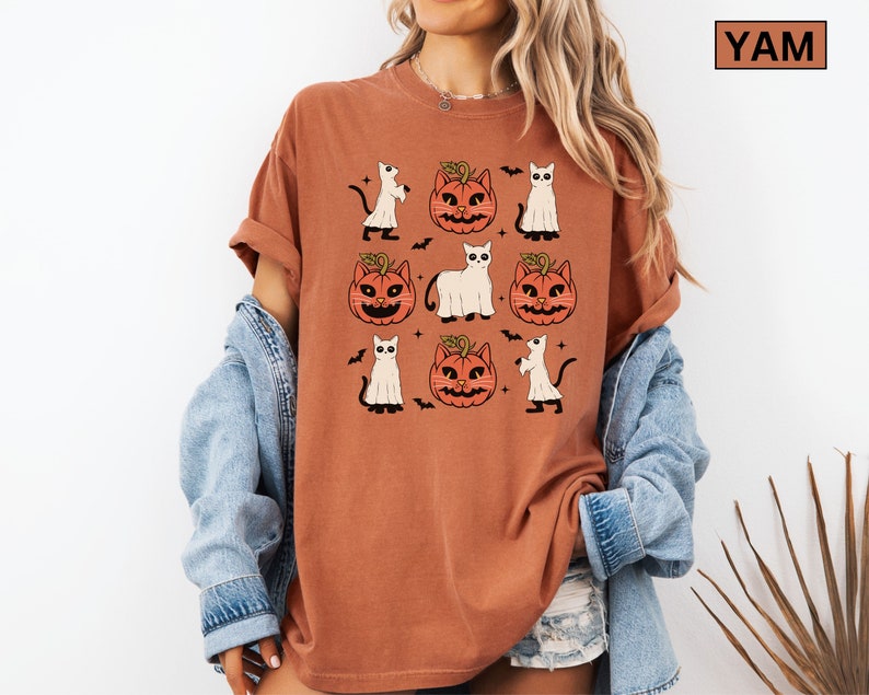 K&ouml;nnte beinhalten: Ein rostorangefarbenes T-Shirt mit einem sich wiederholenden Muster von Halloween-Illustrationen. Das Design umfasst K&uuml;rbislaternen, Geister, Flederm&auml;use und schwarze Katzen. Das Shirt hat hochgekrempelte &Auml;rmel und eine lockere Passform. Der Text "YAM" befindet sich in einem braunen Rechteck.
