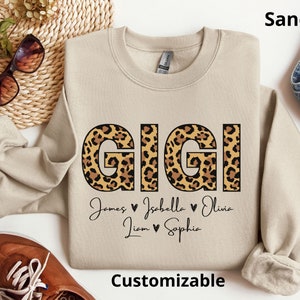 May include: A sand-coloured sweatshirt with a leopard print "GIGI" design and the names "James, Isabella, Olivia, Liam, Sophia" written below. The text "Customizable" is at the bottom of the sweatshirt.