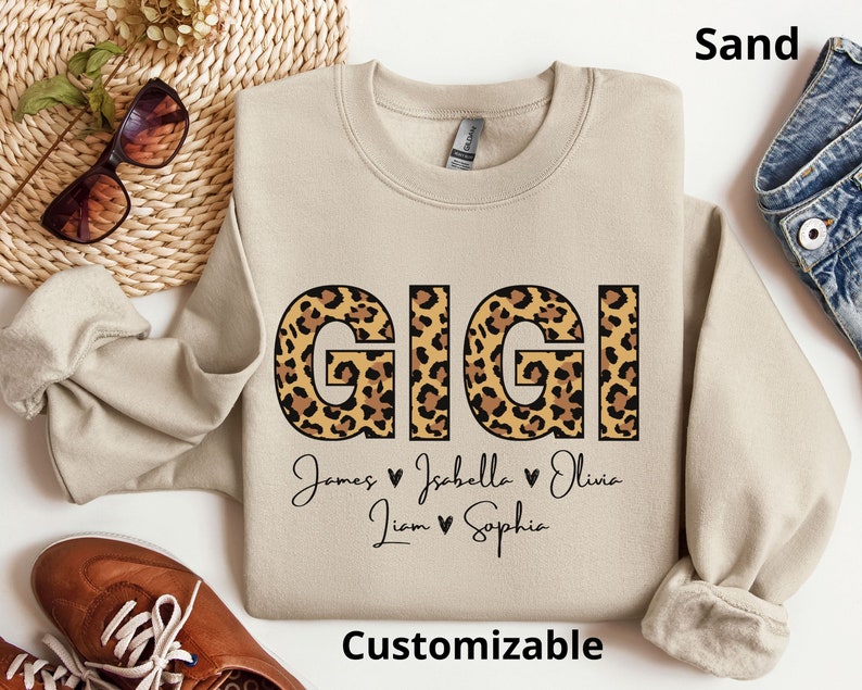 May include: A sand-coloured sweatshirt with a leopard print "GIGI" design and the names "James, Isabella, Olivia, Liam, Sophia" written below. The text "Customizable" is at the bottom of the sweatshirt.