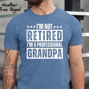 May include: Heather True Royal blue t-shirt with the text "I'M NOT RETIRED I'M A PROFESSIONAL GRANDPA" in white block letters. The shirt has a crew neck and short sleeves. The shirt is made of a soft, comfortable material.