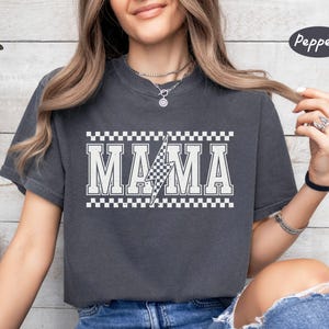May include: Dark grey t-shirt with the word "MAMA" in white block letters, a checkered lightning bolt, and a checkered border. The shirt is a comfortable, casual style.