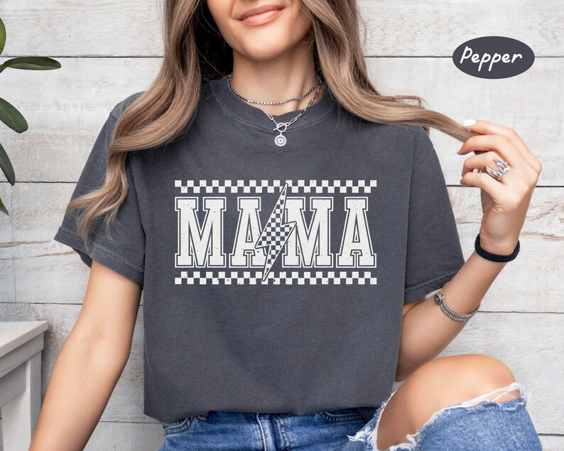 May include: Dark grey t-shirt with the word "MAMA" in white block letters, a checkered lightning bolt, and a checkered border. The shirt is a comfortable, casual style.