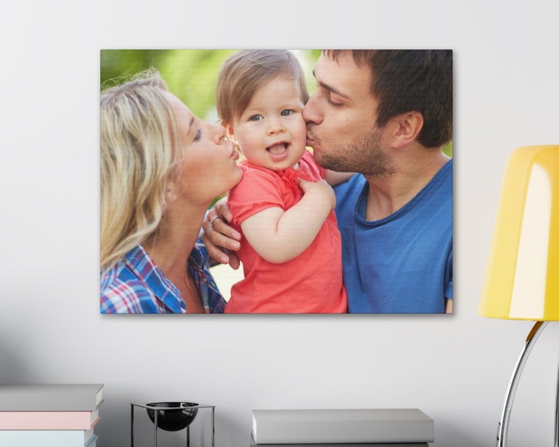 Puede incluir: Impresi&oacute;n en lienzo que presenta un retrato familiar. La imagen muestra a un beb&eacute; sonriendo siendo besado en las mejillas por una mujer y un hombre. El beb&eacute; lleva una camiseta roja. La mujer lleva una camisa a cuadros azul y el hombre una camiseta azul.