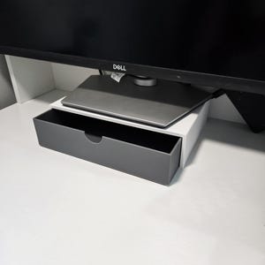 May include: A grey and white monitor stand with a pull-out drawer. The drawer is grey and has a curved handle. A black Dell monitor sits on top of the stand, which is on a white desk.