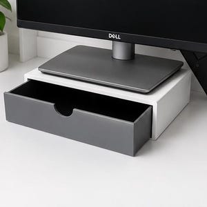May include: A white and grey monitor stand with a pull-out drawer. The grey drawer has a curved cutout for easy opening. A black computer monitor sits on top of the stand.