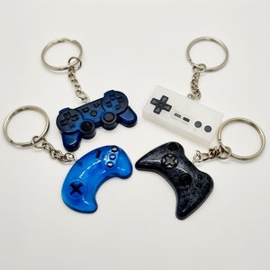 May include: Four different video game controller keychains. One is a blue and black Playstation controller, one is a blue Nintendo controller, one is a black Xbox controller, and one is a white Nintendo Entertainment System controller.