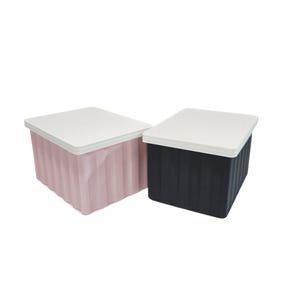 May include: Two storage boxes with white lids. One box is pale pink, and the other is black. Both boxes have a ridged texture on the sides. The boxes are rectangular and appear to be made of plastic.