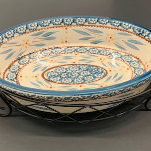 Puede incluir: Una fuente redonda de cerámica para hornear con un diseño floral azul y asas, colocada en un soporte de metal negro. El plato presenta un interior color crema con detalles azules y naranjas. Un artículo de cocina decorativo.