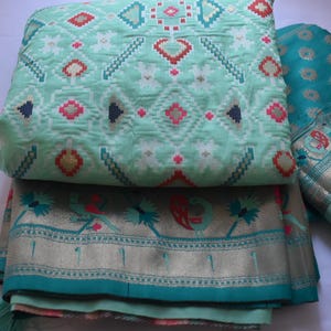May include: A folded mint green fabric featuring a geometric pattern in white, red, and dark blue, paired with a teal fabric border decorated with metallic gold peacock motifs and intricate floral designs along the bottom edge.