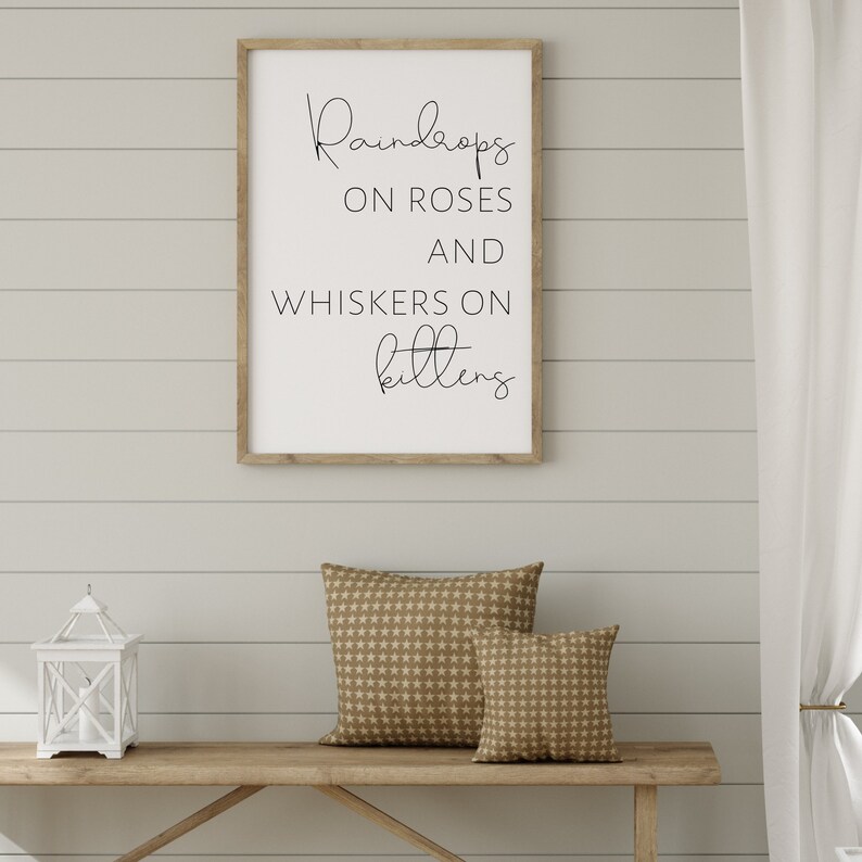May include: A framed print with the words "Raindrops on Roses and Whiskers on Kittens" in black script. Below, a wooden bench holds two patterned pillows and a white lantern. The wall is painted a light gray.