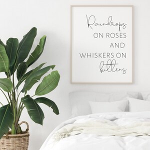 May include: A framed print with the text "Raindrops on Roses and Whiskers on Kittens" in a black script font. The print is in a light wood frame and hangs above a white bed. A large green plant in a woven basket is to the left.