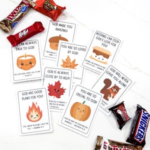 Op de afbeelding: Een assortiment chocoladerepen en kaarten met inspirerende boodschappen en schattige illustraties. De kaarten tonen afbeeldingen van een kaars, eikel, blad, s'more, esdoornblad, eekhoorn, vuur en pompoen. De chocoladerepen zijn onder andere Kit Kat, Reese's, Twix en Snickers.