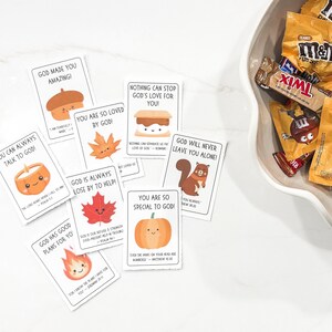 Op de afbeelding: Een verzameling kleine, witte kaarten met kleurrijke illustraties en tekst, waaronder een pompoen, een eikel en een esdoornblad. De kaarten bevatten inspirerende boodschappen en bijbelverzen. Een kom met snoepjes in fun size staat op de achtergrond.