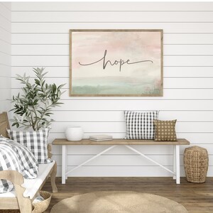 May include: A watercolor painting with the word "hope" in cursive script on a light pink and green background. The painting is framed and hanging on a white shiplap wall above a wooden bench with pillows.