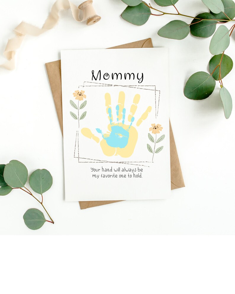 May include: A white card with the words "Mommy" in black script. A yellow handprint with a blue handprint inside is in the center of the card. The text "Your hand will always be my favorite one to hold." is below the handprint.