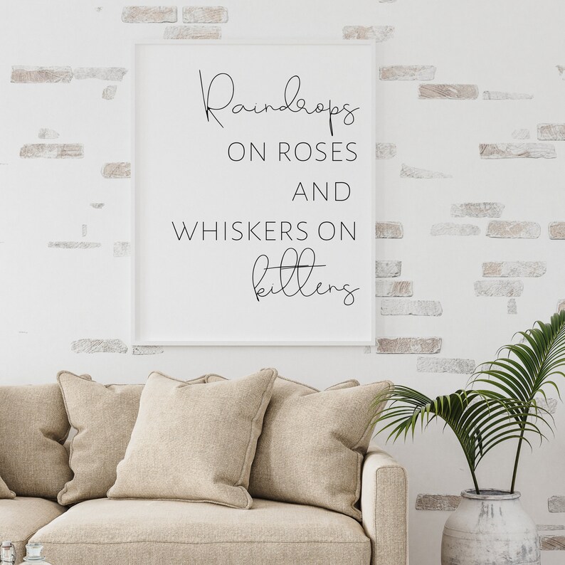 May include: A framed print with the words "Raindrops on Roses and Whiskers on Kittens" in a stylish script. The print is displayed on a white brick wall above a beige sofa with matching pillows. A decorative vase with a green plant is on the right.