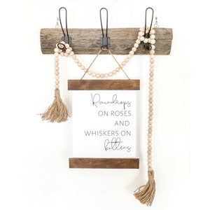 May include: A rustic wooden wall decor piece with three metal hooks. A white sign with the text "Raindrops on Roses and Whiskers on Kittens" is suspended below. Wooden beads and tassels add a decorative touch.
