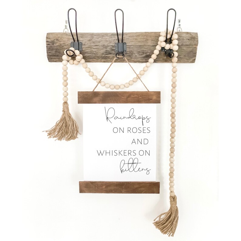 May include: A rustic wooden wall decor piece with three metal hooks. A white sign with the text "Raindrops on Roses and Whiskers on Kittens" is suspended below. Wooden beads and tassels add a decorative touch.