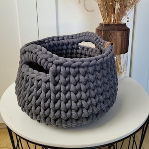 May include: A gray knitted storage basket with two leather handles. The basket is sitting on a white table with a black metal base.