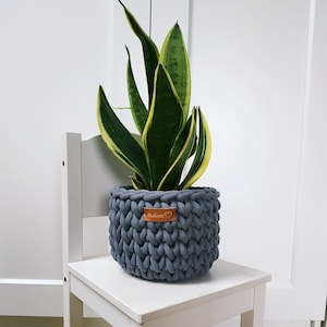 May include: A gray crocheted plant pot with a brown leather tag that says "Babern" on it. A snake plant is in the pot, and the pot is sitting on a white wooden stool.