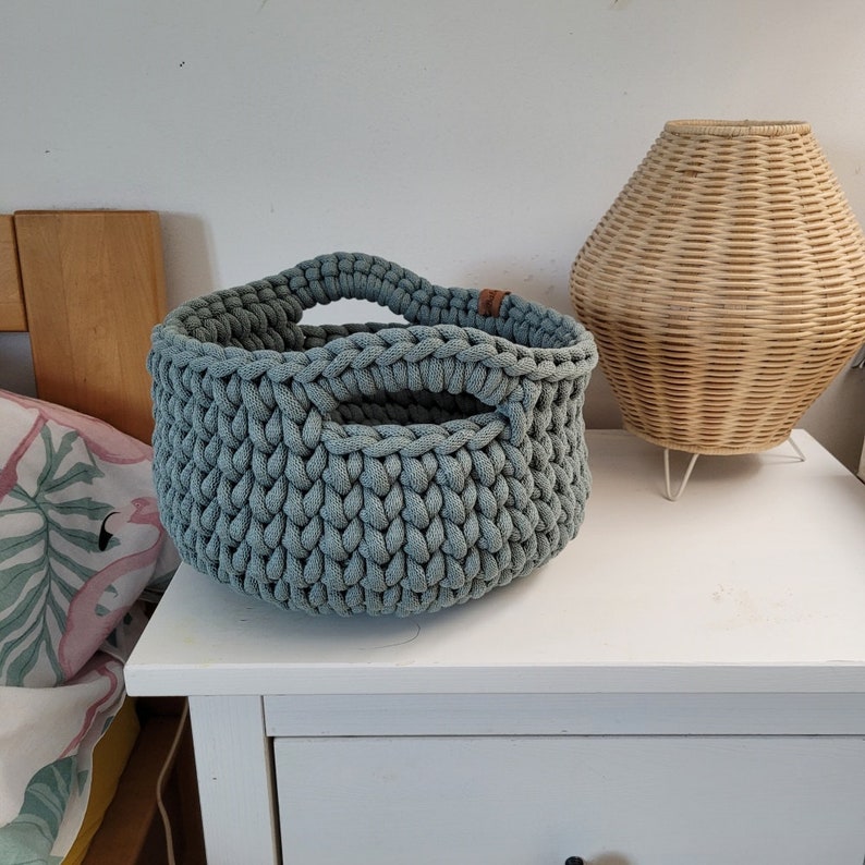 May include: A light blue crocheted storage basket with a handle. The basket is sitting on a white nightstand with a drawer.