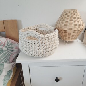 May include: A white crocheted basket with two handles sits on a white nightstand with a drawer. The basket is made of thick rope and has a textured surface.