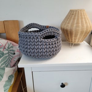 May include: A gray crocheted storage basket with a handle. The basket is sitting on a white nightstand with a drawer.
