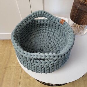 May include: A light blue crocheted basket with a single handle. The basket is made of thick cotton rope and has a round shape.