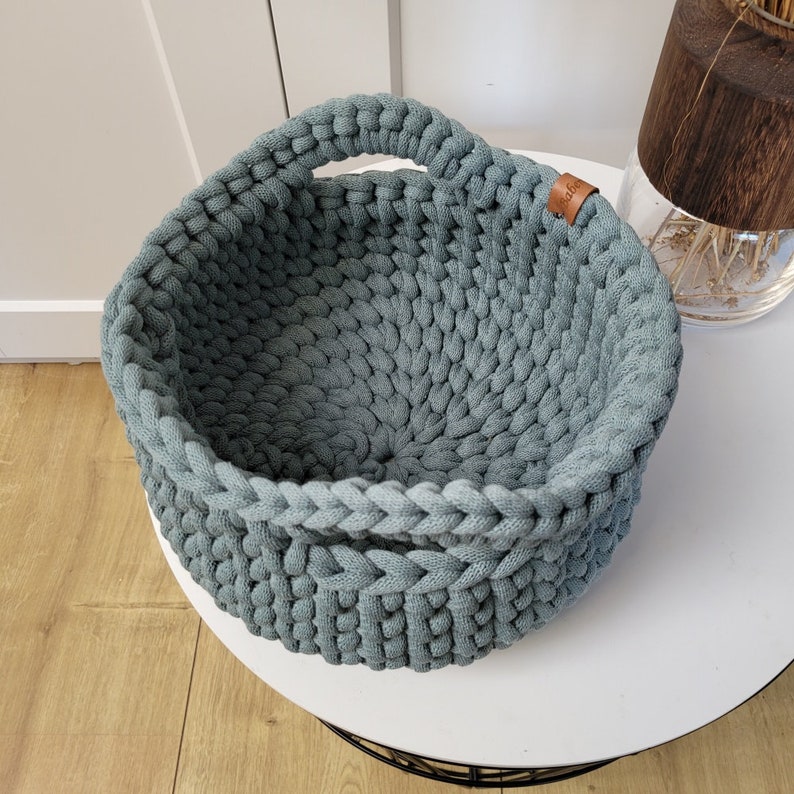 May include: A light blue crocheted basket with a single handle. The basket is made of thick cotton rope and has a round shape.