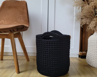 May include: A black, cylindrical, hand-knitted storage basket with two sturdy handles. The basket is made of thick, textured yarn and has a simple, modern design. The basket is approximately 12 inches in diameter and 14 inches tall.