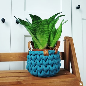 Könnte beinhalten: Ein türkisfarbener gehäkelter Pflanzenhänger mit braunen Lederriemen hält eine Topfpflanze mit Schlangenpflanze. Der Pflanzenhänger steht auf einem Holzregal.