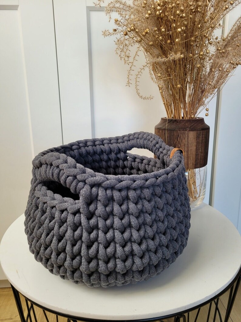 May include: A gray knitted storage basket with two leather handles. The basket is sitting on a white table with a black metal base.