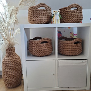 May include: A white shelf unit with four shelves and a drawer. The shelves are filled with brown crocheted baskets. There is a vase on the bottom shelf, also crocheted.