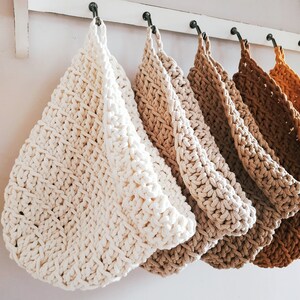 May include: Five crocheted hanging baskets in various shades of beige and brown, hanging from a white shelf with metal hooks.