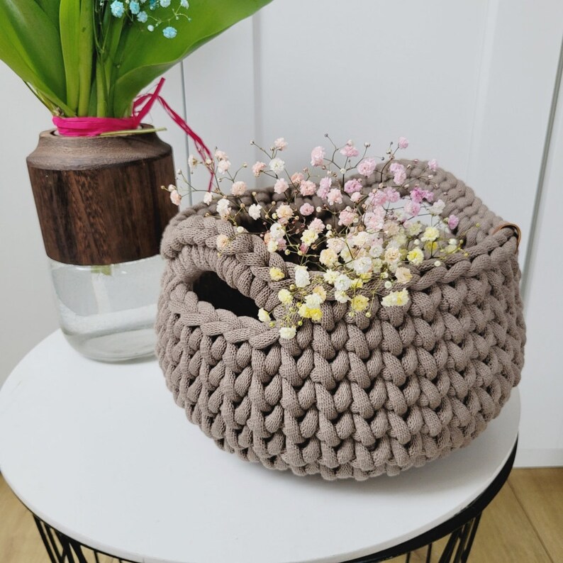 May include: A brown crocheted basket with a small opening at the top. The basket is filled with a bouquet of small pink and white flowers.