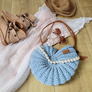 Könnte beinhalten: Eine hellblaue gehäkelte Tasche in Form einer Muschel mit einer Reihe weißer Perlen um die Öffnung. Die Tasche hat einen braunen Lederriemen und ein kleines braunes Lederetikett mit dem Text "Made with Love" darauf.