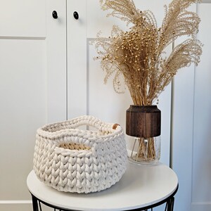May include: A white crocheted storage basket with a wooden handle. The basket is sitting on a white table with a vase of dried flowers in the background.